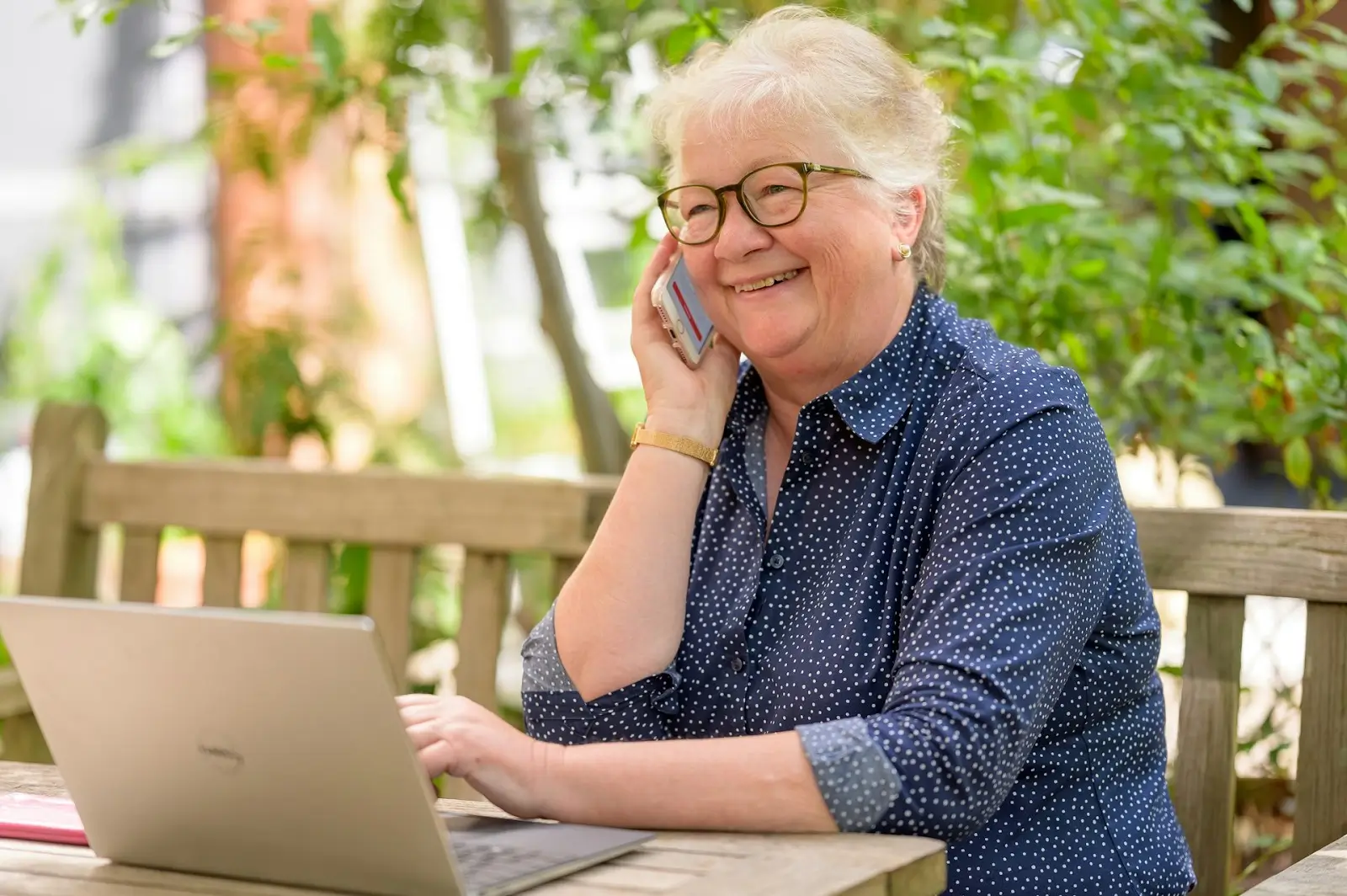 Smiling elderly woman wearing glasses and a blue polka-dot shirt talking on a phone while using a laptop outdoors.