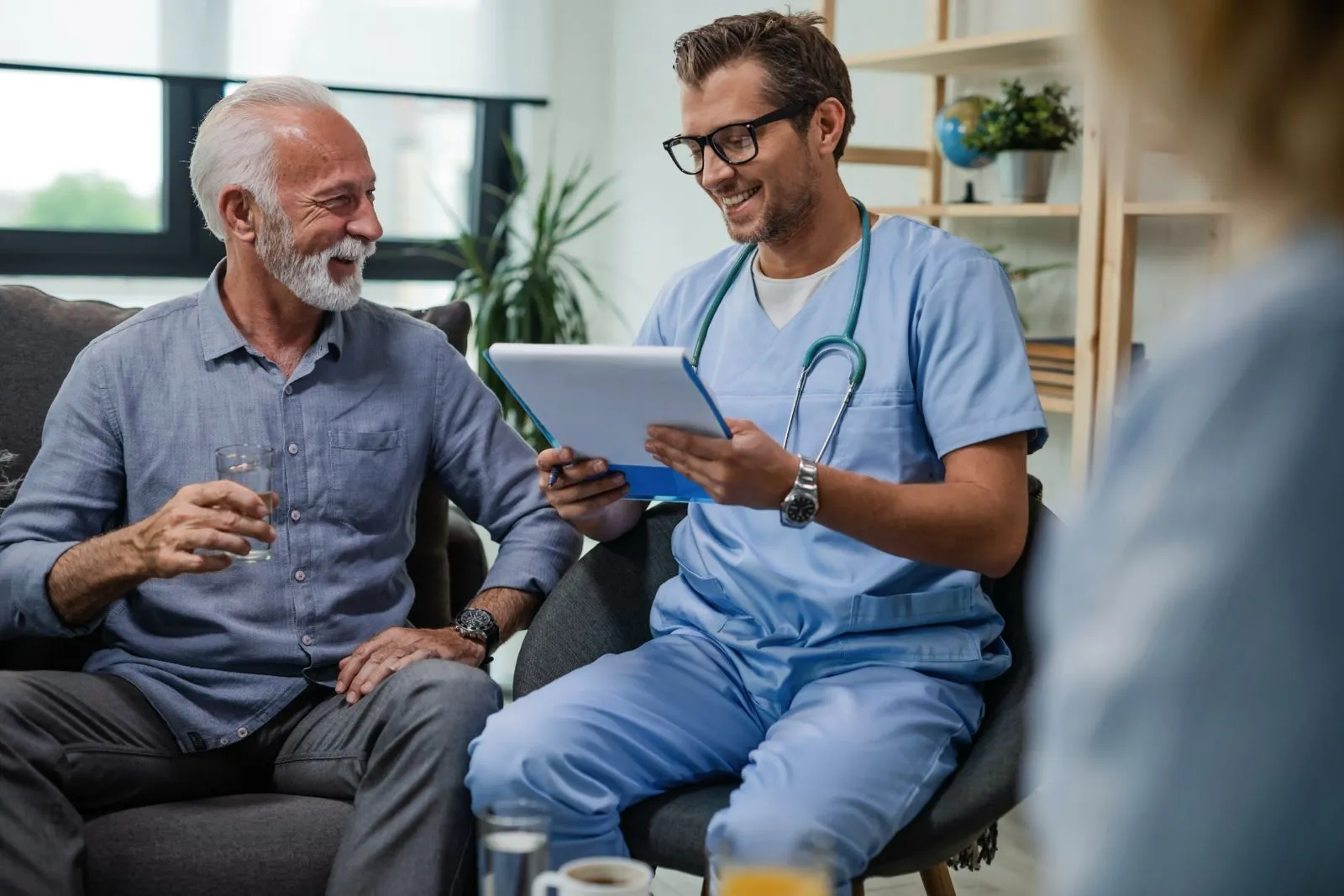 Smiling elderly man holding a glass of water talking with a male healthcare professional in scrubs reviewing a clipboard.