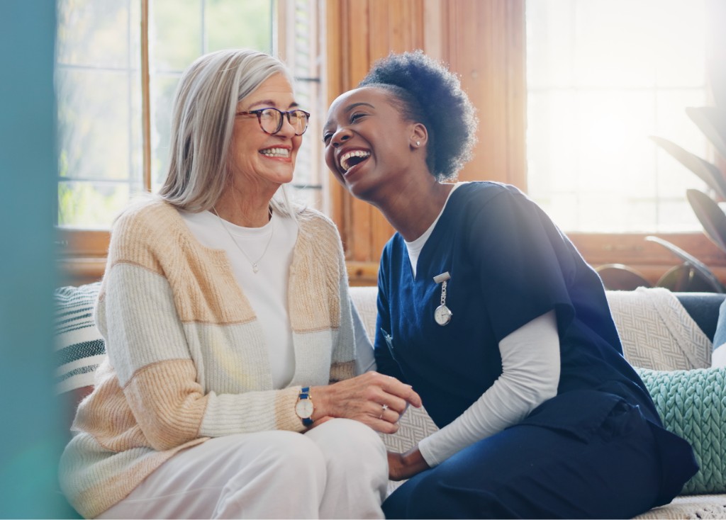 A smiling elderly woman on a sofa with a young female nurse in blue scrubs laughing joyfully beside her.