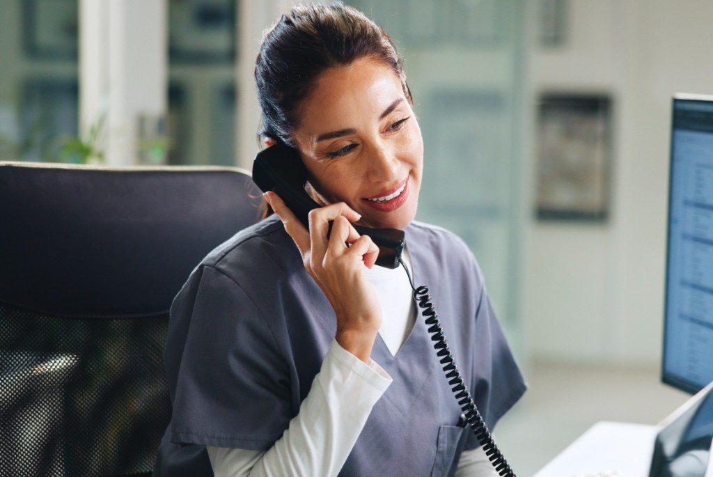 Smiling practice staff member in grey scrubs on the phone at a desk with a computer in a bright modern office.